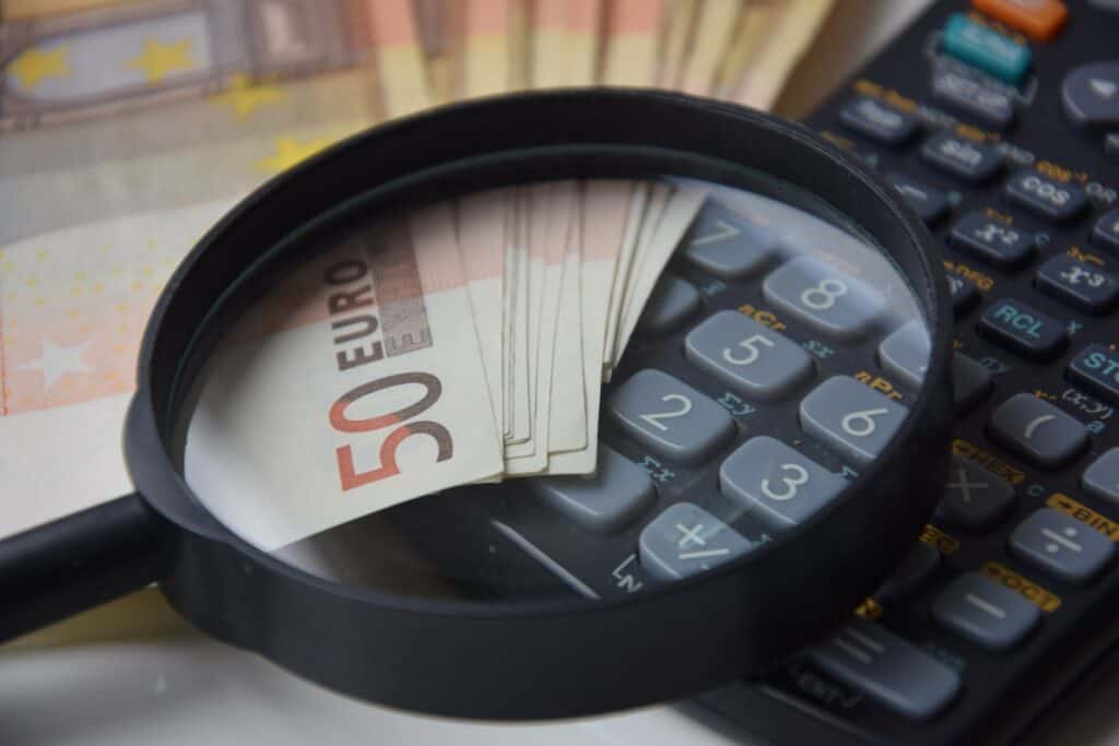 Man sieht einen Bündel 50 Euro Scheine und einen Taschenrechner. Darauf liegt eine Lupe, die das 50 Euro Symbol des Geldscheines vergrößert
