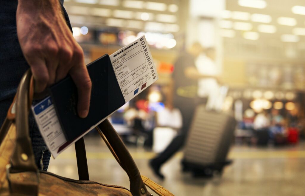 Man sieht eine Hand mit Reisepass und Flugticket (Bordkarte). Im Hintergrund ist ein Flughafenterminal erkennbar.