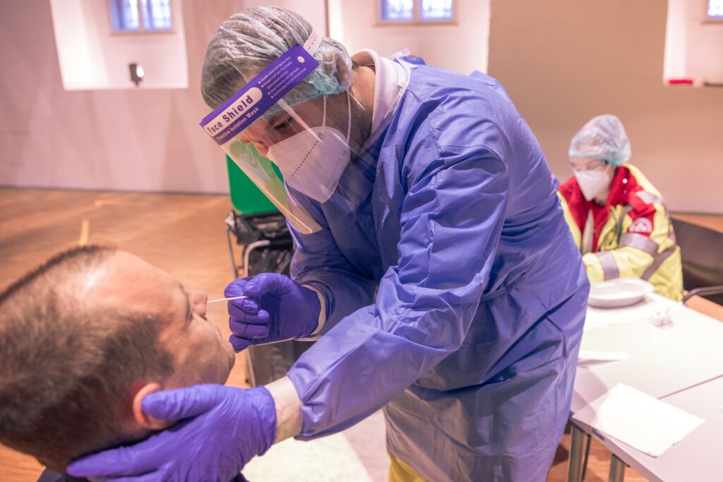 Es ist eine Person zu sehen, die einen Mund-Nasen Abstrich per Wattestäbchen durch die Nase bekommt. Zwei medizinische Angestellte in Schutzkleidung sind zu sehen.