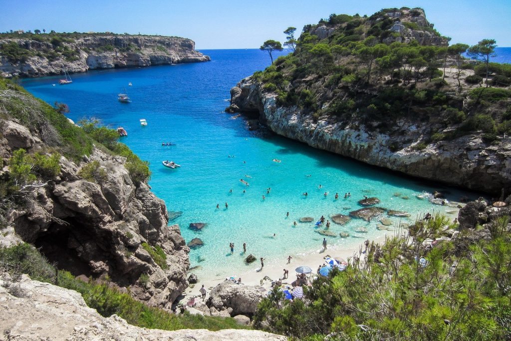 Es ist eine Bucht im Mittelmeer mit Strand und Urlaubern vor einem blauen Himmel zu erkennen.
