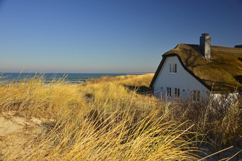 Es ist ein Haus mit Strohdach zu erkennen. Im Hintergrund sieht man das Meer, vorne sind Gräser und Sträucher zu sehen.