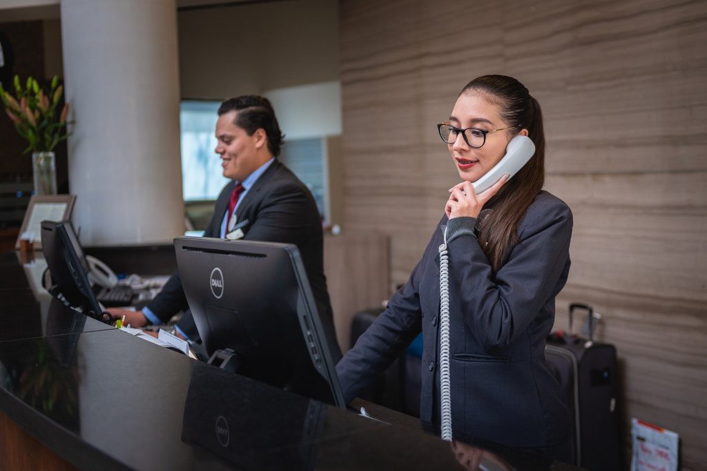 Es ist die Rezeption eines Hotels zu erkennen. Eine Frau telefoniert, ein anderer Mitarbeiter links schaut in einen Computerbildschirm.