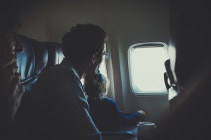 Es ist ein Mann mit einem Kind auf dem Schoß im Flugzeug zu sehen. Beide Personen schauen aus dem Fenster.