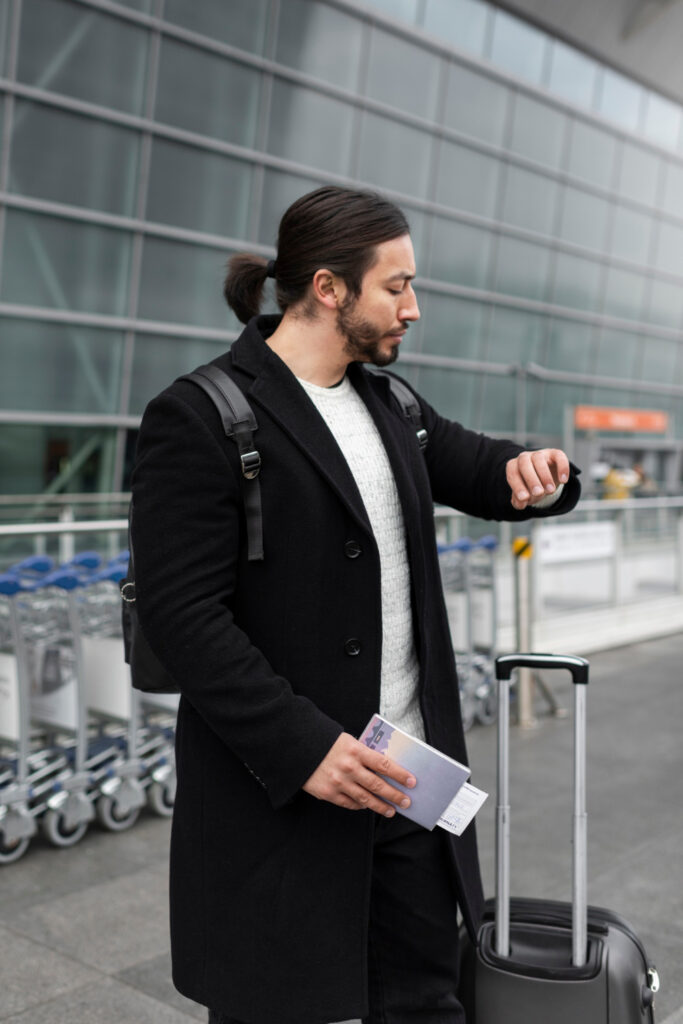 Mann mit Koffer und Flugtickets schaut skeptisch auf seine Armbanduhr; steht vor einem Flughafen. 