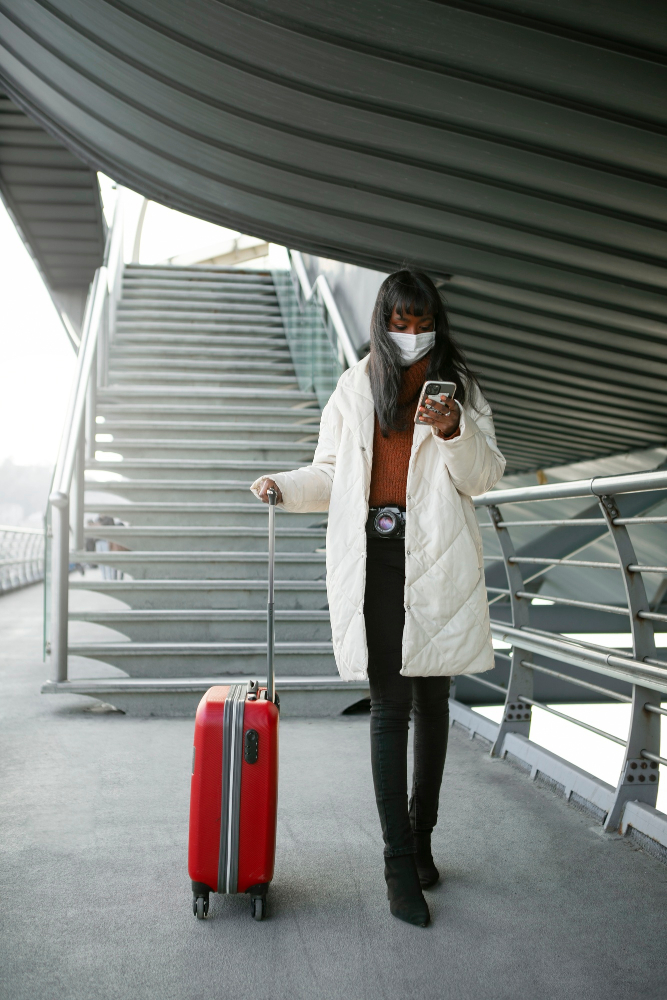 Eine Frau mit schwarzen Haaren und Mundschutz checkt ihr Smartphone und zieht ihren Koffer.