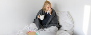 Kranke blonde Frau mit Tasse in der Hand schnäuzt sich die Nase.