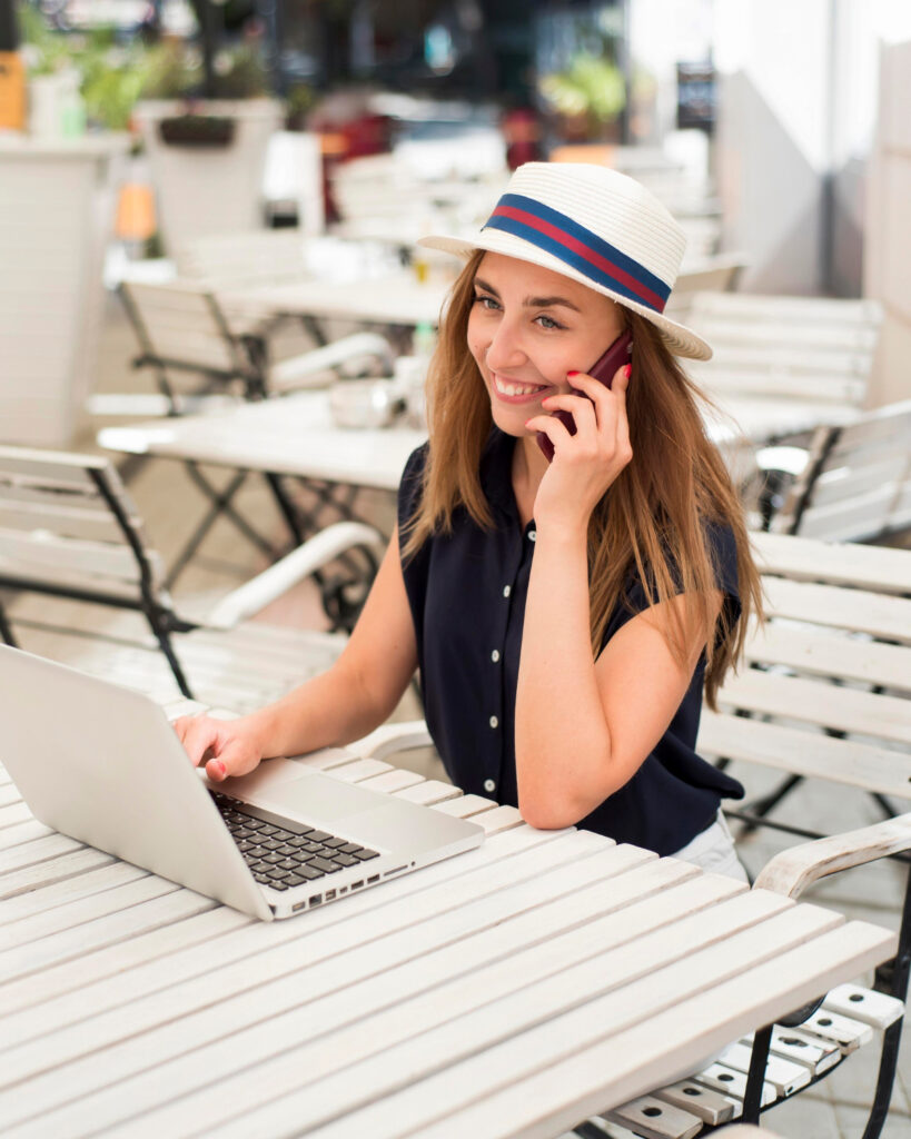 Frau mit Sommerhut arbeitet draussen am Laptop und telefoniert.