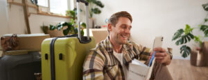 Portrait of smiling tourist man sits with suitcase holds smartphone passport and tickets goes on holiday